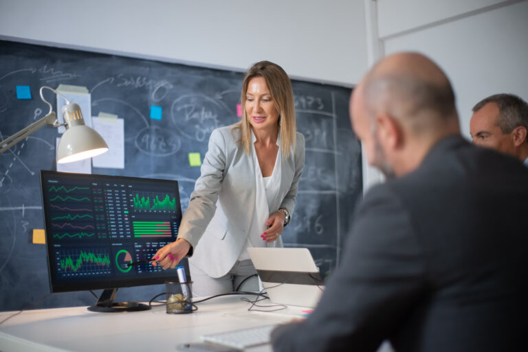 Executiva vestindo terno cinza apresentando gráficos e dados em um monitor para uma equipe de gestores, destacando como a IA para reduzir custos e aumentar a eficiência operacional otimiza decisões estratégicas.