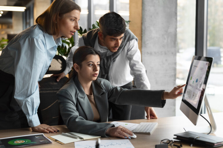 Três profissionais analisam dados em um computador durante uma reunião de trabalho. Uma mulher aponta para a tela enquanto os outros dois colegas observam atentamente, demonstrando colaboração para otimizar a eficiência operacional.