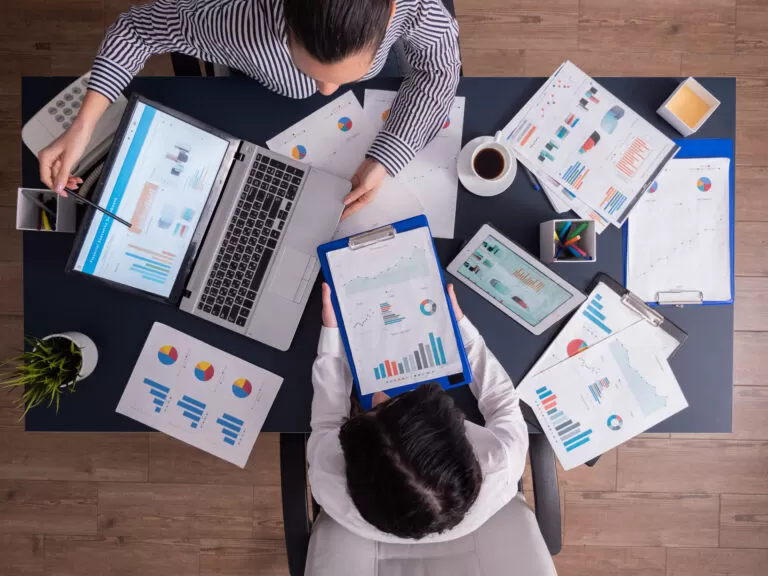 Imagem vista de cima mostrando duas pessoas em uma mesa de trabalho rodeadas por gráficos impressos, notebook, tablet e documentos. Elas analisam dados com apoio visual e digital, representando o uso de software personalizado corporativo para tomada de decisões estratégicas.