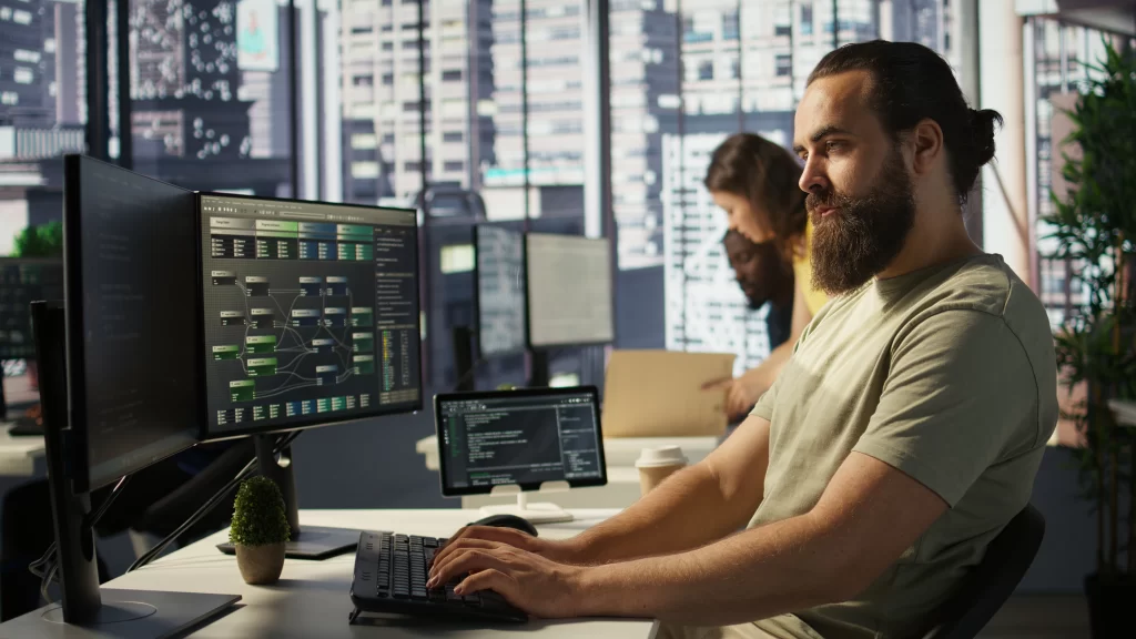 Profissional de TI programando em dois monitores com fluxos de dados e código, enquanto equipe trabalha ao fundo em escritório moderno — simbolizando colaboração e eficiência no outsourcing de TI.